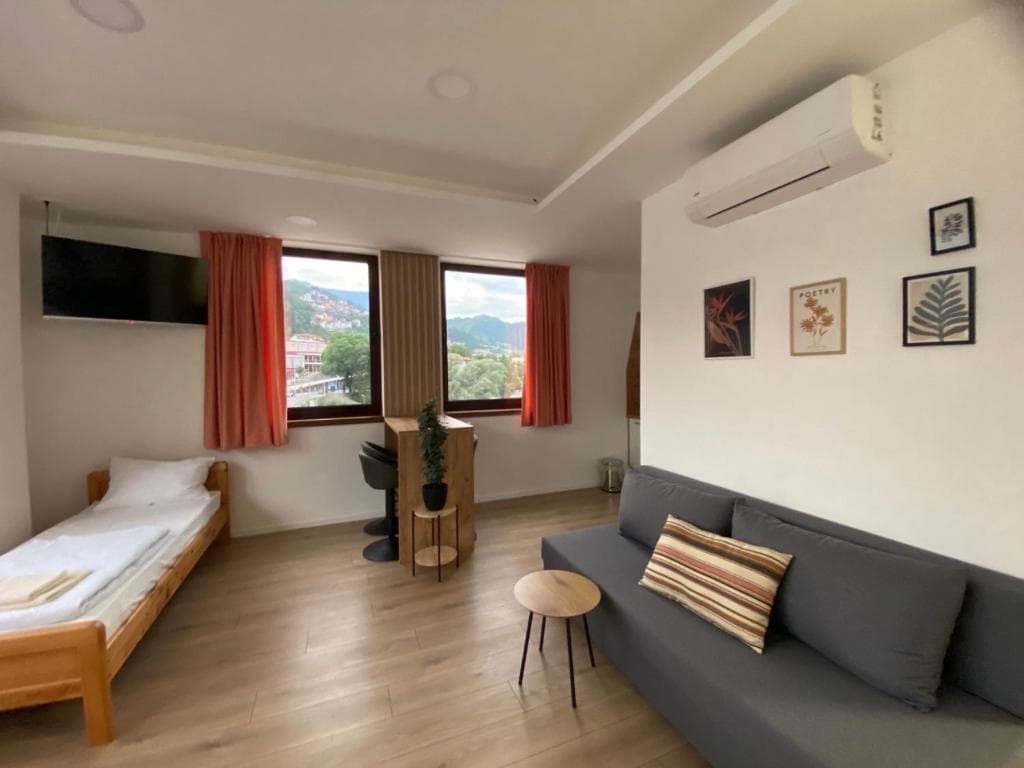 Open-plan studio with sleeping area, high table by the windows, and grey sofa under framed art.